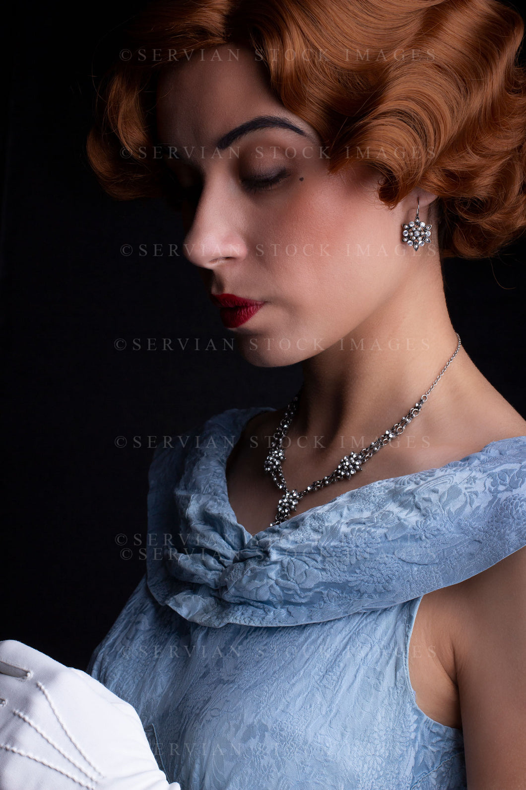 1930s woman wearing a blue vintage dress, long white gloves, and a diamante necklace against a black backdrop.(Sarah 0235)