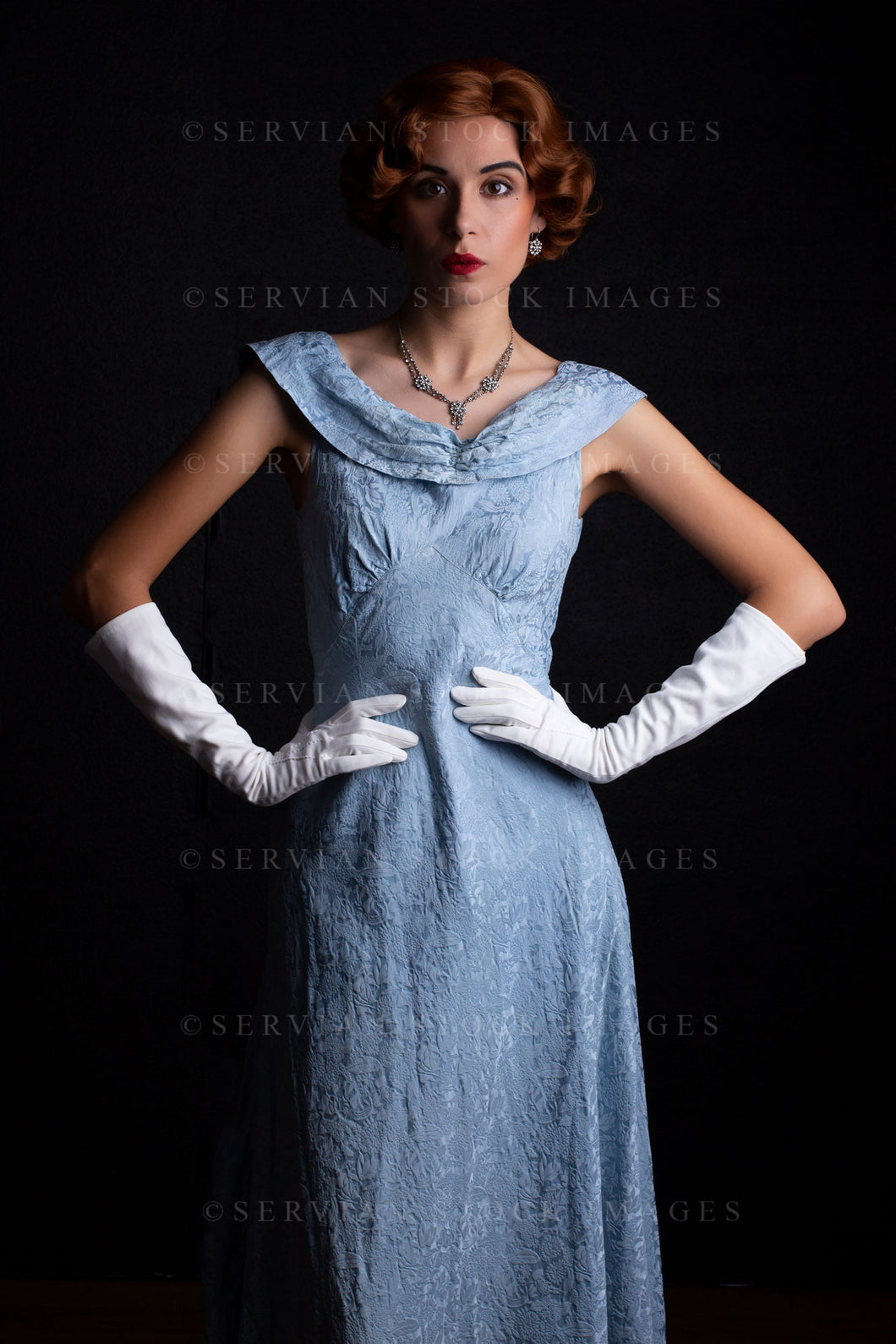 1930s woman wearing a blue vintage dress, long white gloves, and a diamante necklace against a black backdrop.(Sarah 0220)