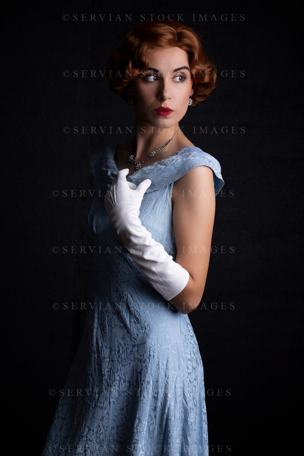 1930s woman wearing a blue vintage dress, long white gloves, and a diamante necklace against a black backdrop. (Sarah 0233)