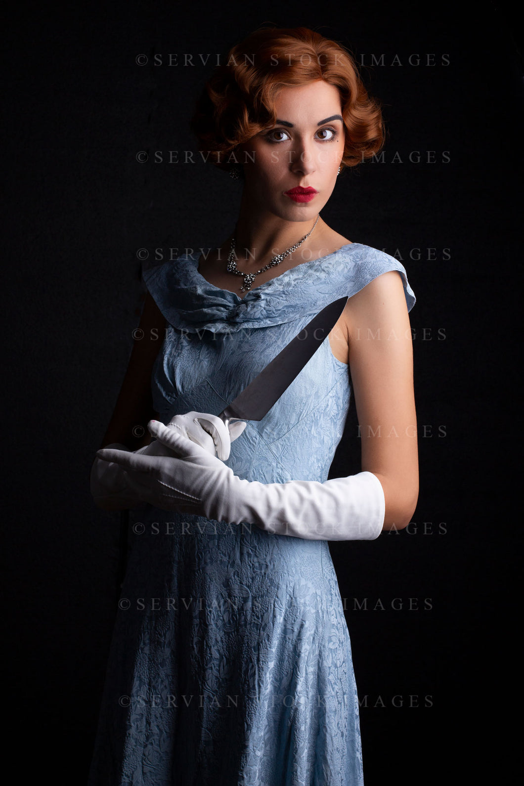 1930s woman wearing a blue vintage dress, long white gloves, and a diamante necklace. She's holding a large kitchen knife against a black backdrop.(Sarah 0254)