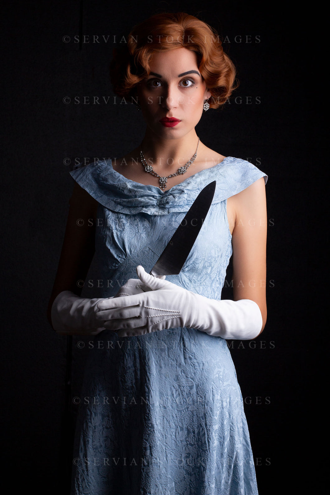 1930s woman wearing a blue vintage dress, long white gloves, and a diamante necklace. She's holding a large kitchen knife against a black backdrop.(Sarah 0257)