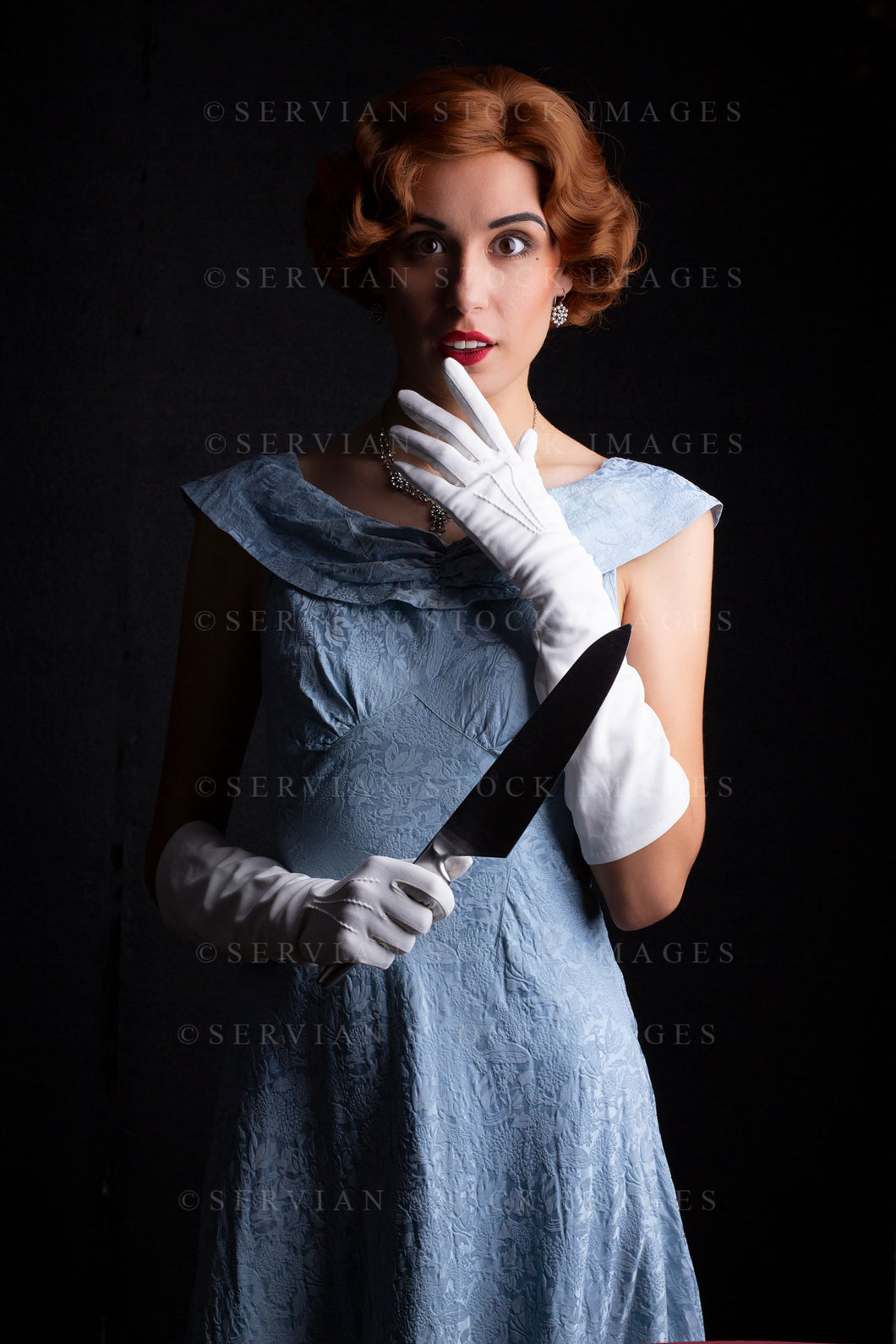 1930s woman wearing a blue vintage dress, long white gloves, and a diamante necklace. She's holding a large kitchen knife against a black backdrop.(Sarah 0259)