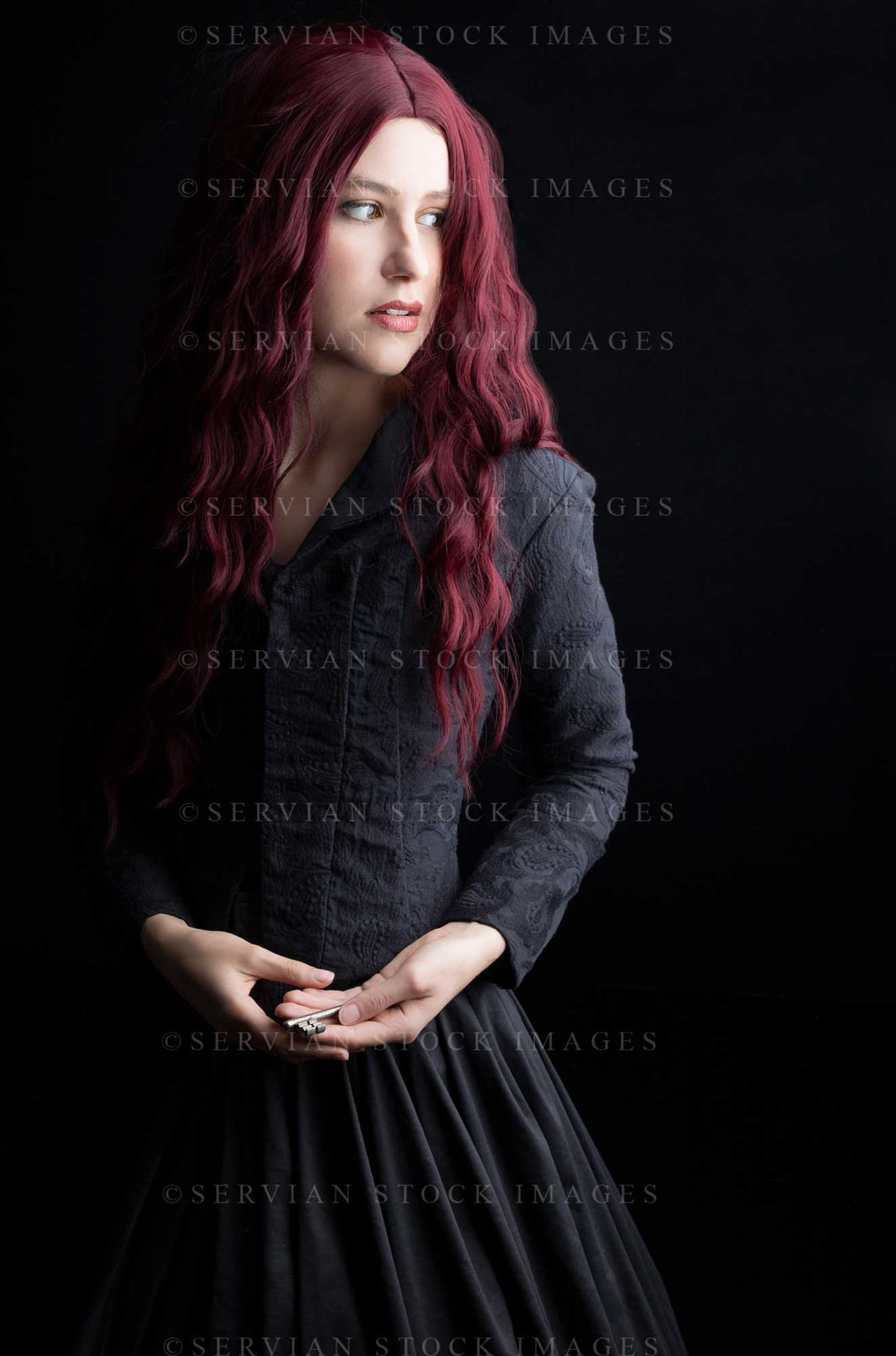 Victorian woman with long red hair wearing a black bodice and skirt against a black backdrop and holding an antique key. She could also be a witch or a ghost. (Amalia 0846)